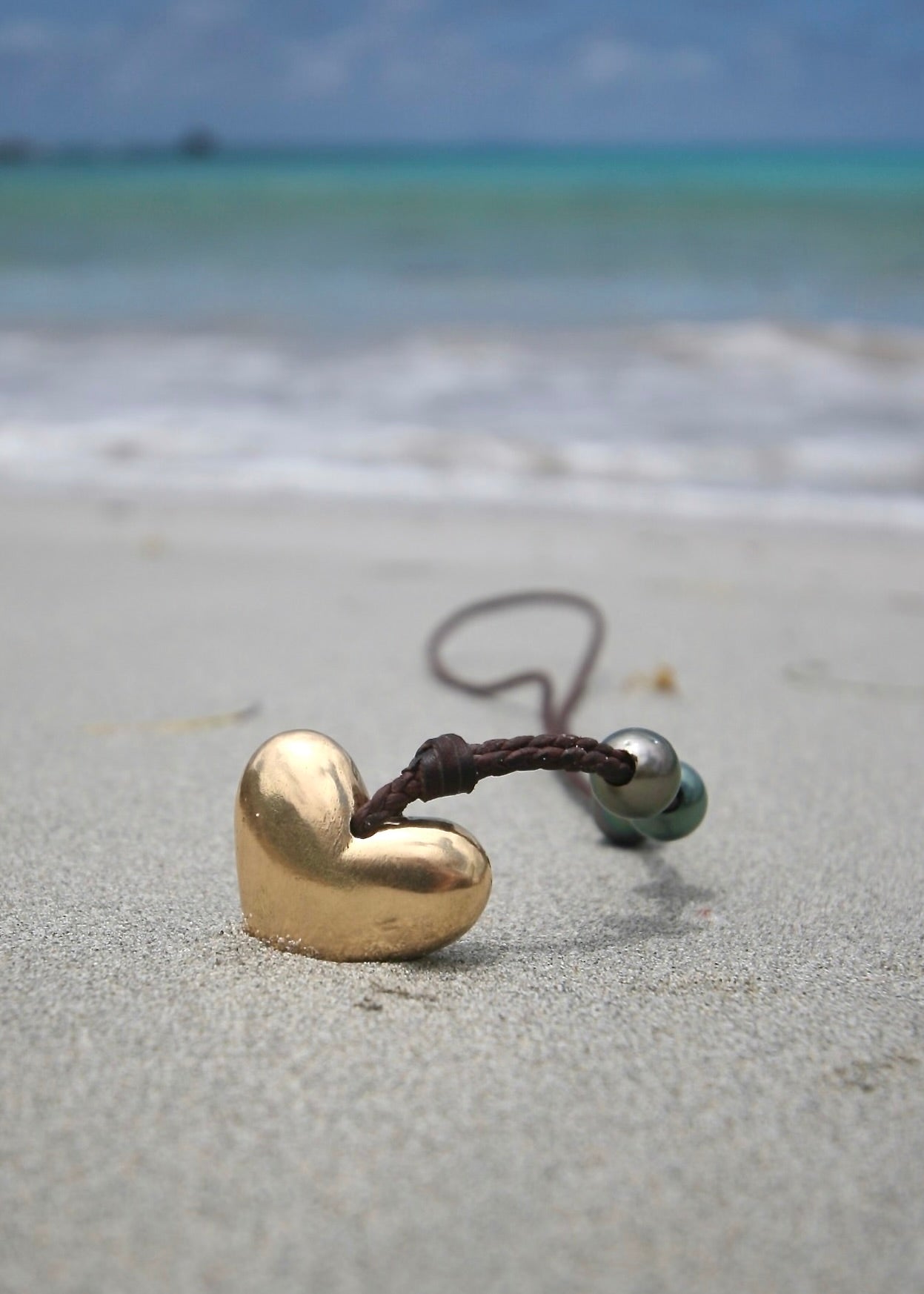 Solid gold heart and Tahitian pearl leather necklace on sand, Caribbean sea