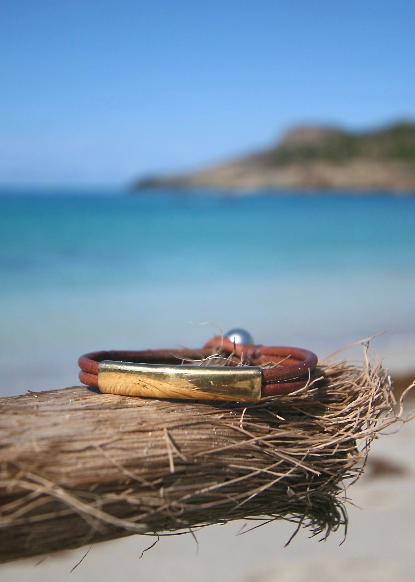 Trésors de St Barth 18K solid gold bar bracelet on kangaroo leather resting dispatch on driftwood with ocean behind
