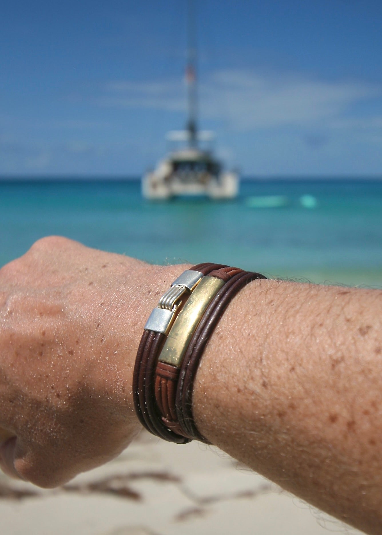 Hand-sculpted 18K gold bar bracelet worn on wrist with turquoise Caribbean sea and sailboat in background