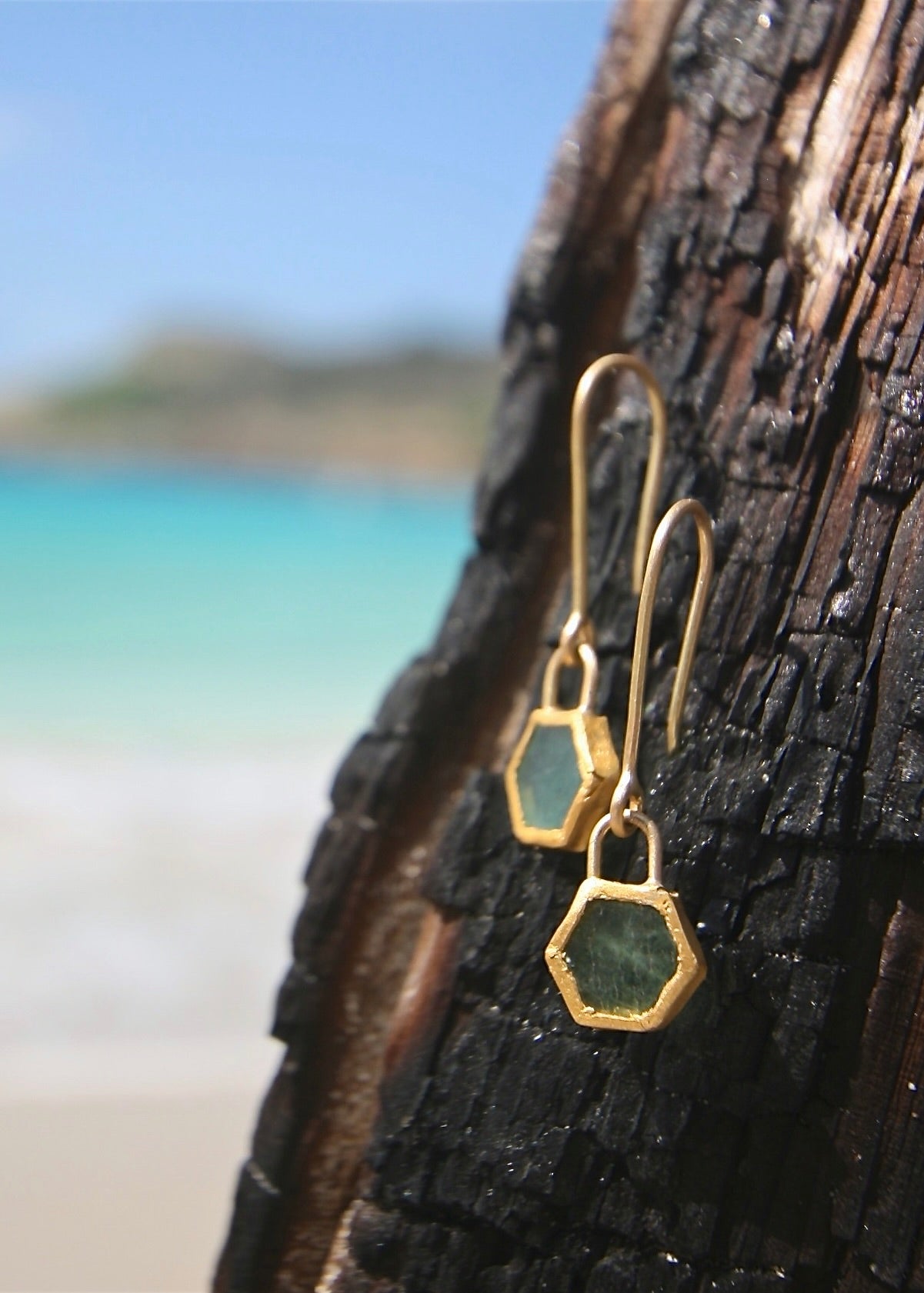 Trésors de St Barth raw emerald hexagonal gold earrings on dark wood, blue sea
