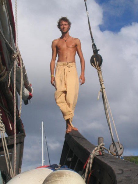 Ted Deltour standing barefoot on a sailboat rigging, shirtless under the sky