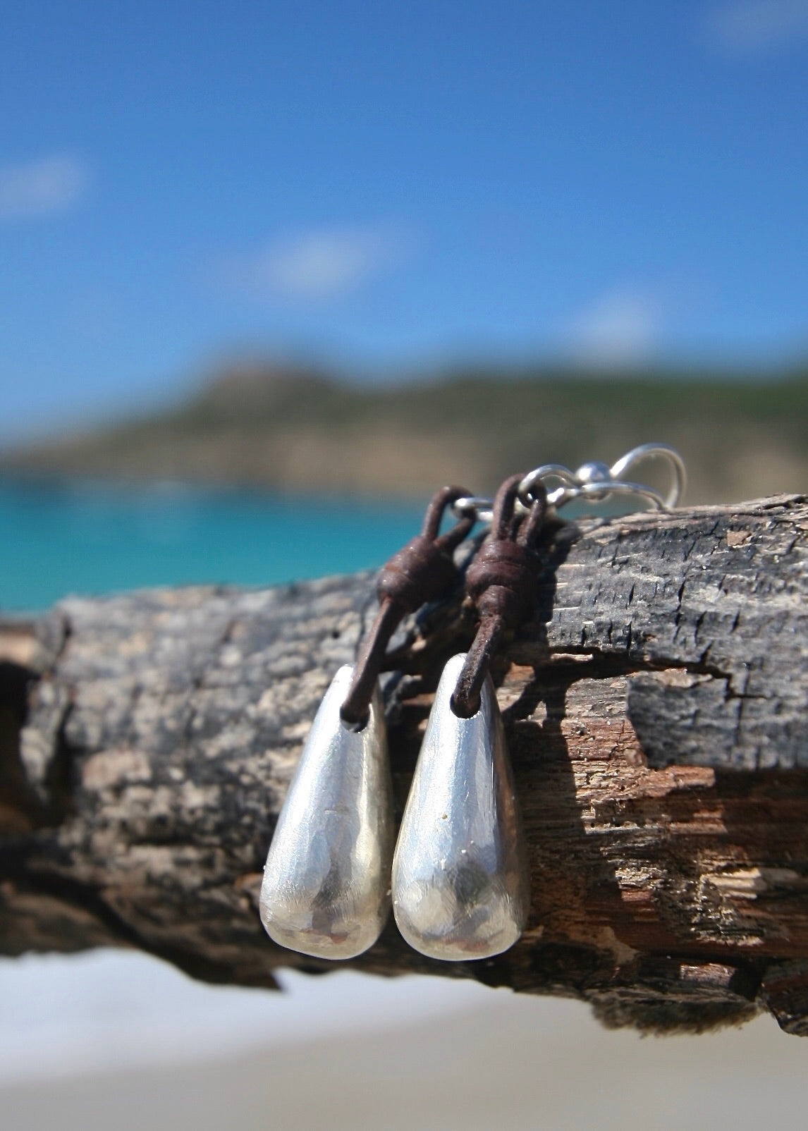 Hand-sculpted silver drop leather earrings on driftwood, Caribbean turquoise sea