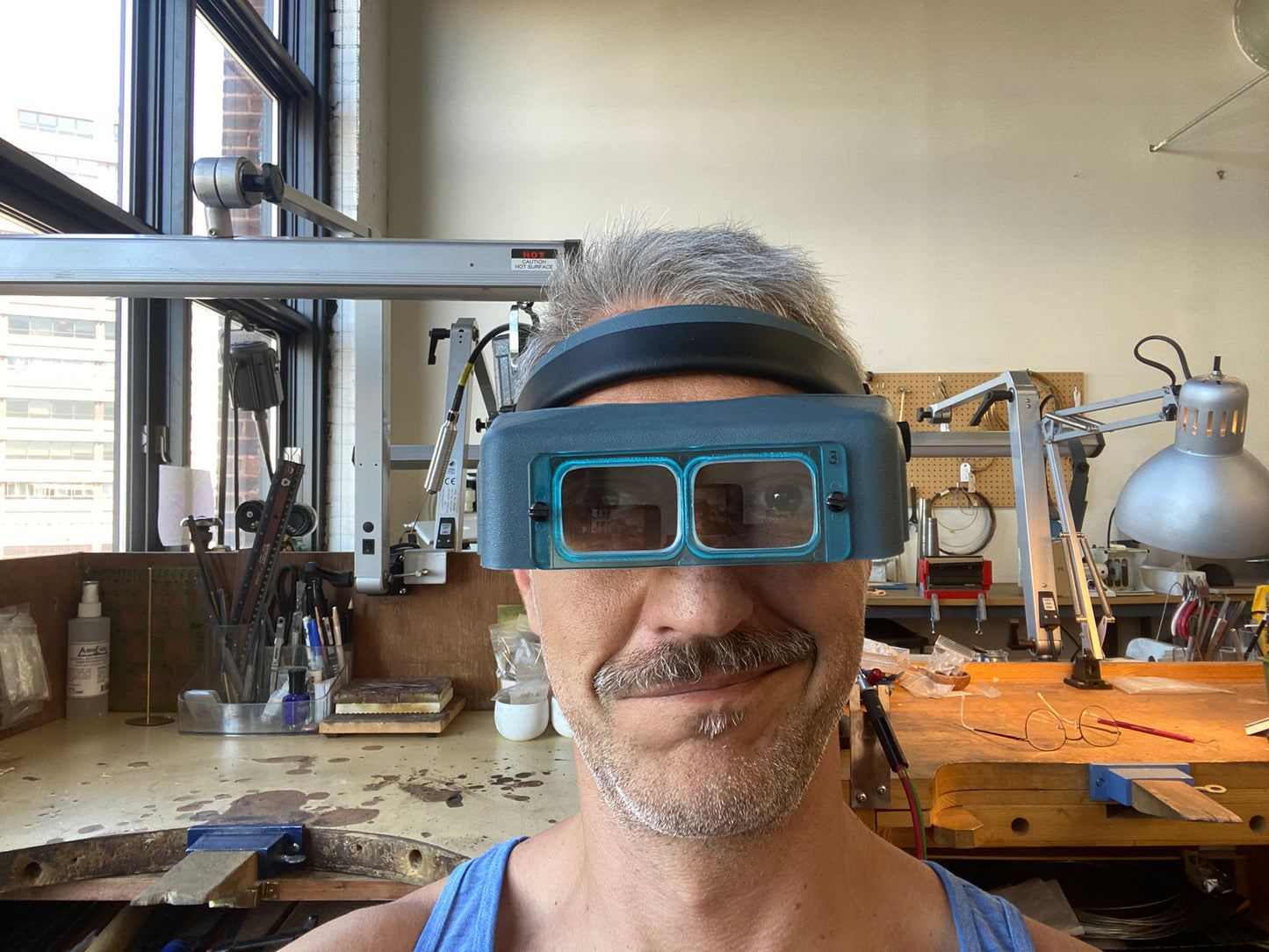 A man wearing a pair of blue and black dual-lens magnifying glasses in a workshop setting.