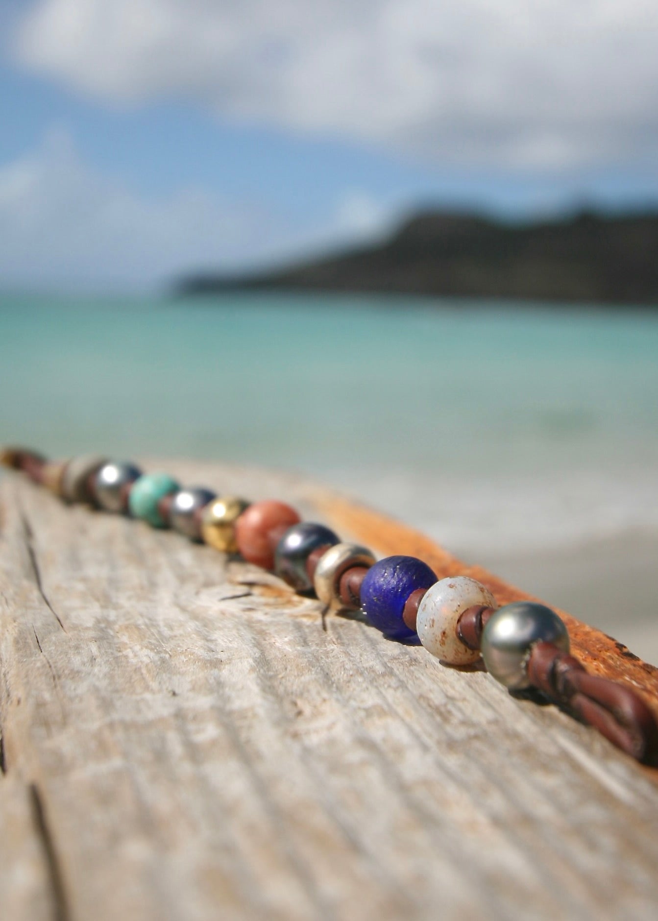 Bracelet stretched along driftwood, all beads visible in a line, St. Barth sea and hillside
Treasure bracelet with Tahitian pearls, 18k gold beads, turquoise, lapis lazuli and rare ancient beads on kangaroo leather, displayed on driftwood with St. Barth sea — handmade jewelry by Trésors de St Barth.