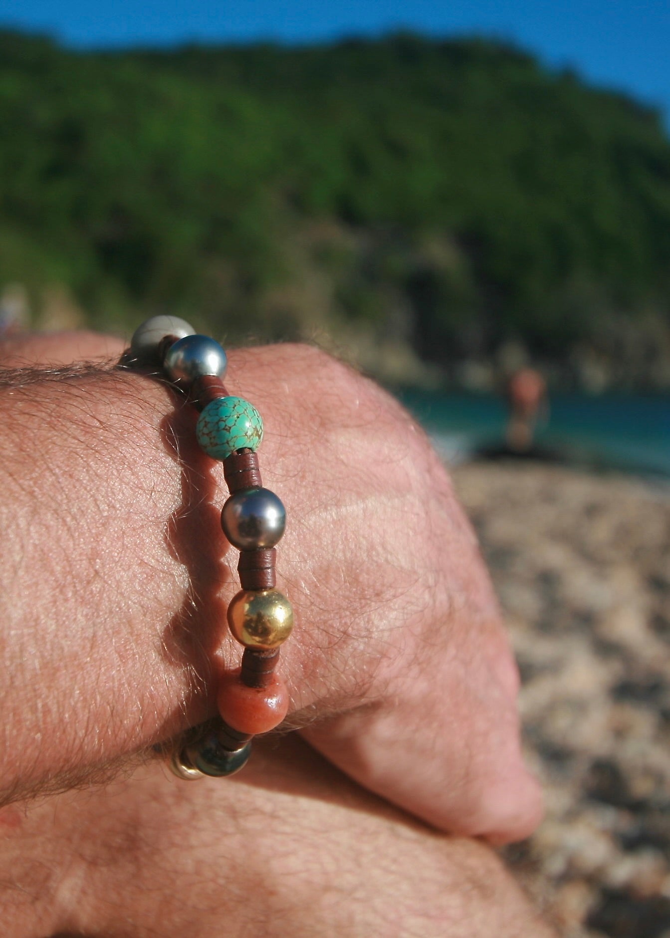 Lifestyle wrist shot on man's wrist, St. Barth beach and green hillside in background
Treasure bracelet with Tahitian pearls, 18k gold and rare beads on kangaroo leather worn on man's wrist at a St. Barth beach — unisex luxury boho bracelet by Trésors de St Barth.
