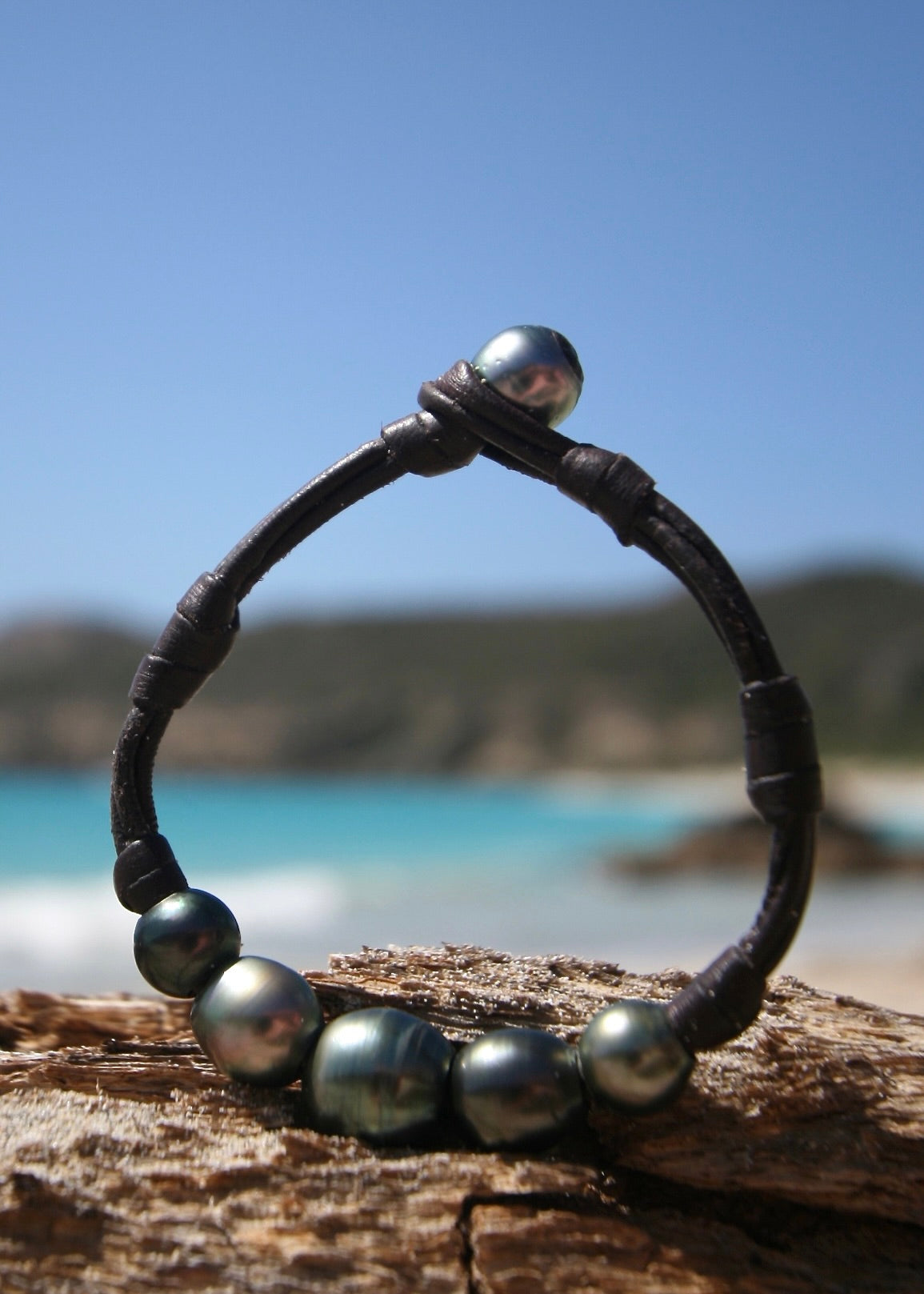 Dark Tahitian pearl leather bracelet on driftwood, Caribbean turquoise sea
