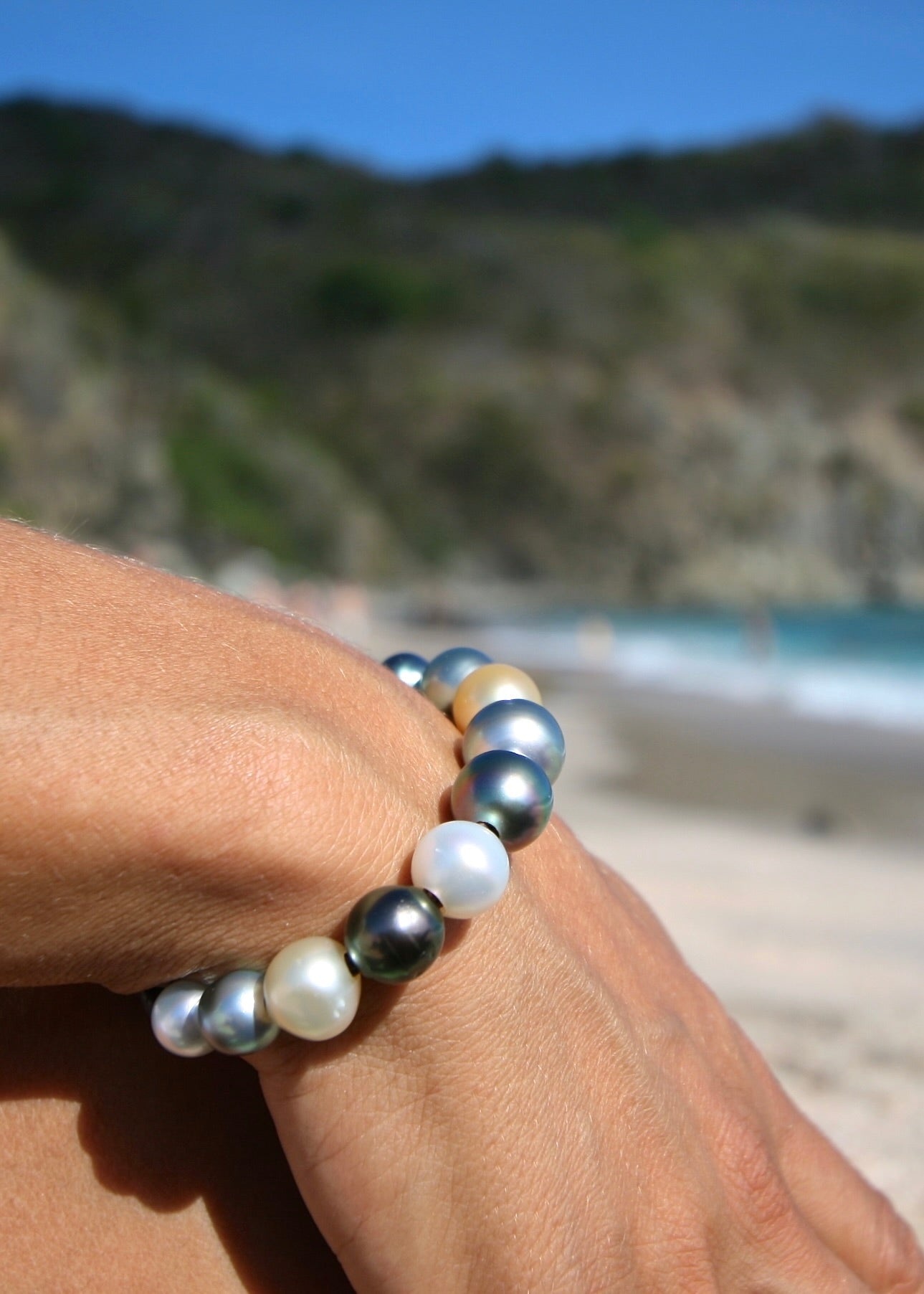Multicolor Tahitian and Australian pearl gradient bracelet worn on wrist with ocean and hillside behind