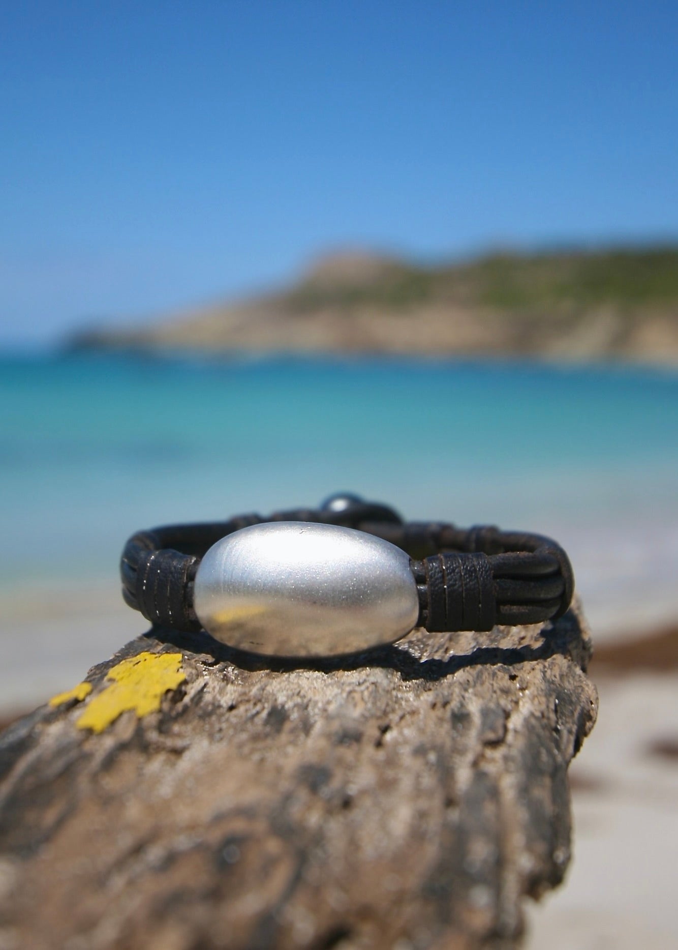 Hand-sculpted sterling silver pebble on kangaroo leather bracelet displayed on driftwood at Caribbean beach