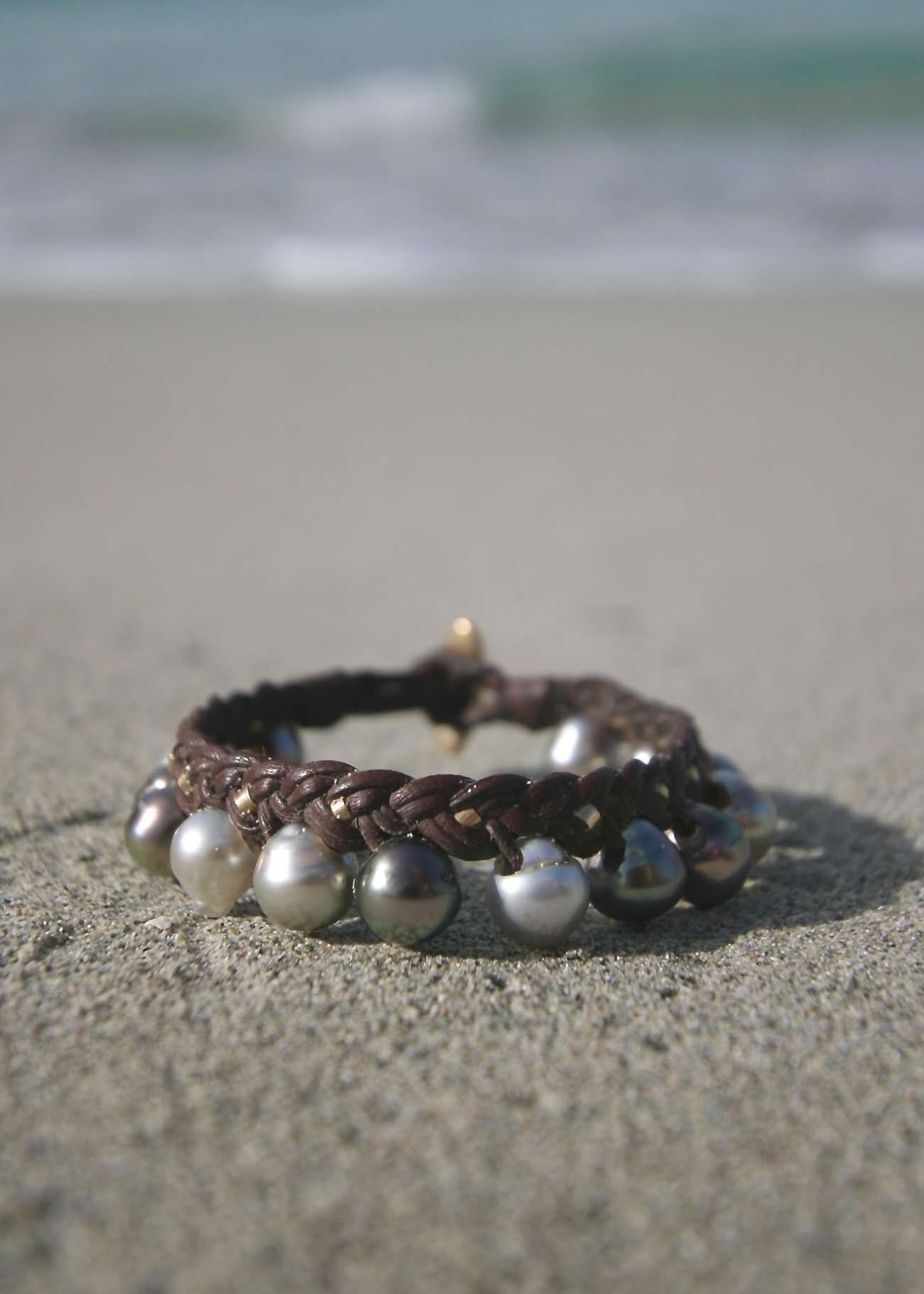 Tahitian pearl and 18K gold braided kangaroo leather bracelet circular view on white sand with ocean waves
