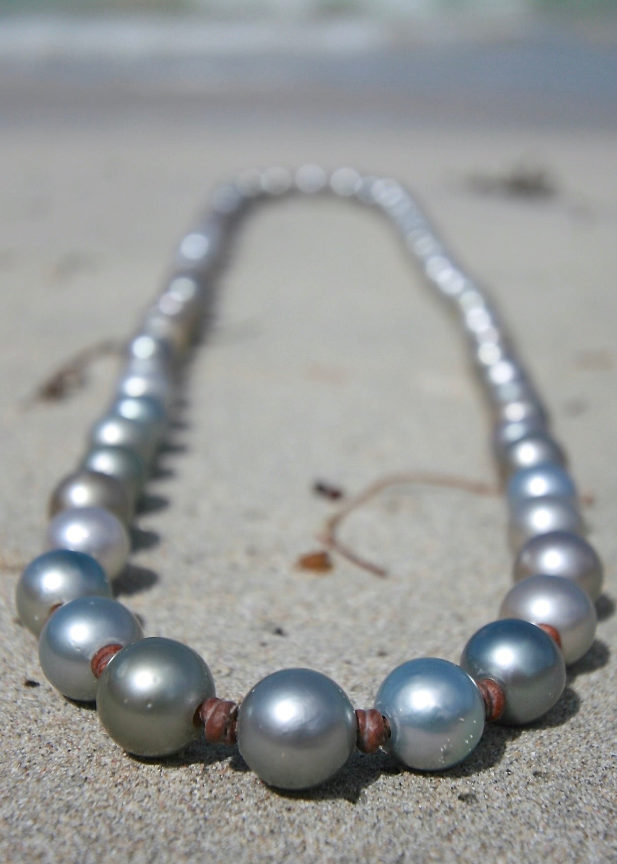 Detail of 12mm round Tahitian silver pearls on kangaroo leather necklace laid on white sand showing size